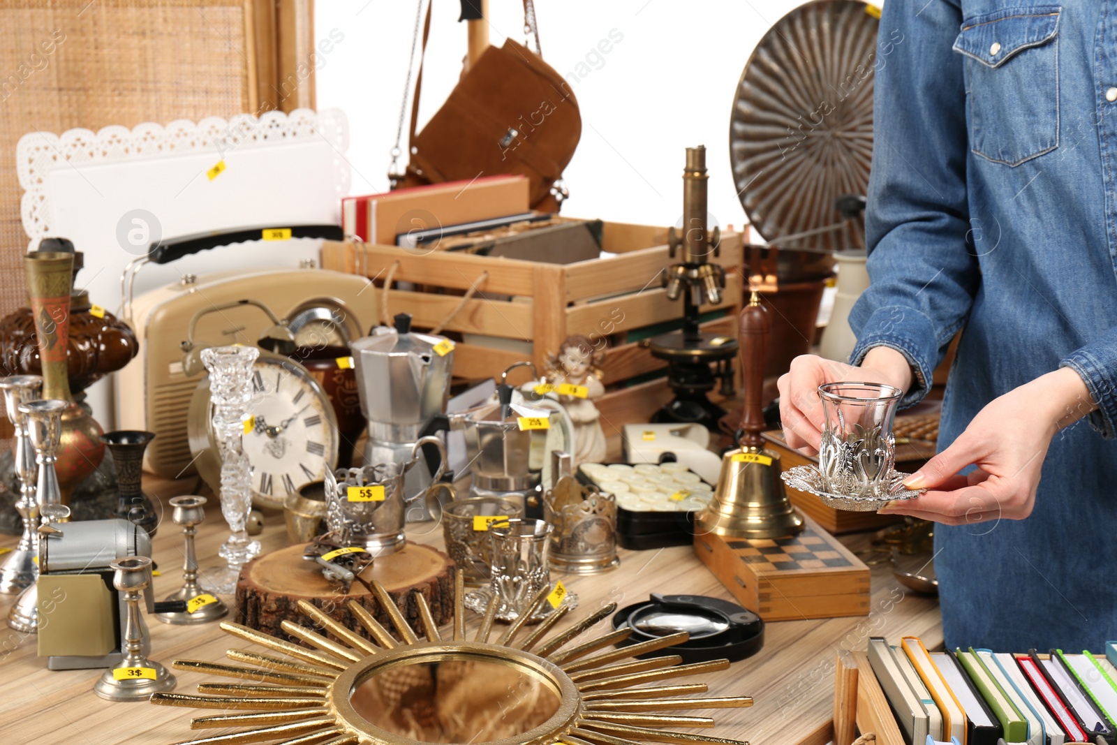 Photo of Woman holding glass cup with saucer near table with different stuff indoors, closeup. Garage sale