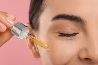 Young woman applying serum onto her face on pink background, closeup