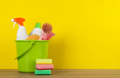 Bucket with different cleaning supplies on wooden floor near yellow wall. Space for text
