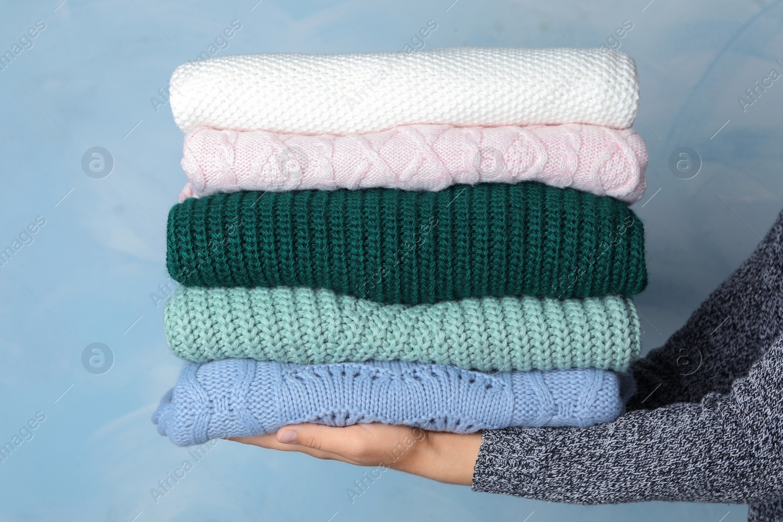 Photo of Woman holding stack of warm clothes on light blue background, closeup. Autumn season