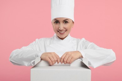 Happy professional confectioner in uniform holding cake box on pink background