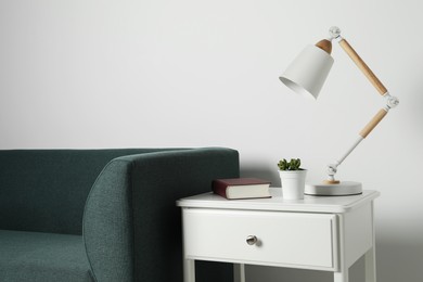 Photo of Stylish lamp, houseplant with book on side table and soft sofa near white wall. Interior design
