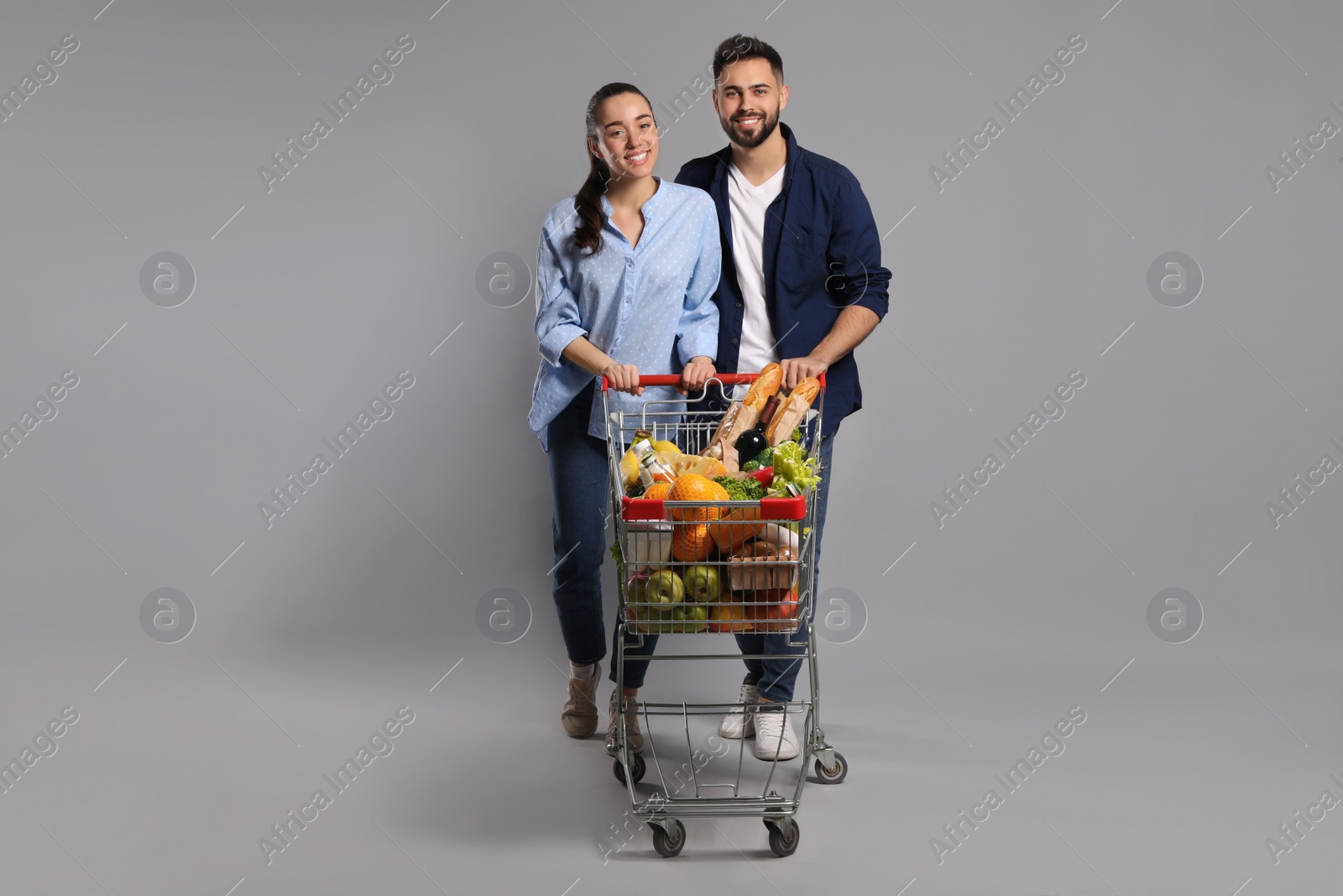 Photo of Happy couple with shopping cart full of groceries on light grey background