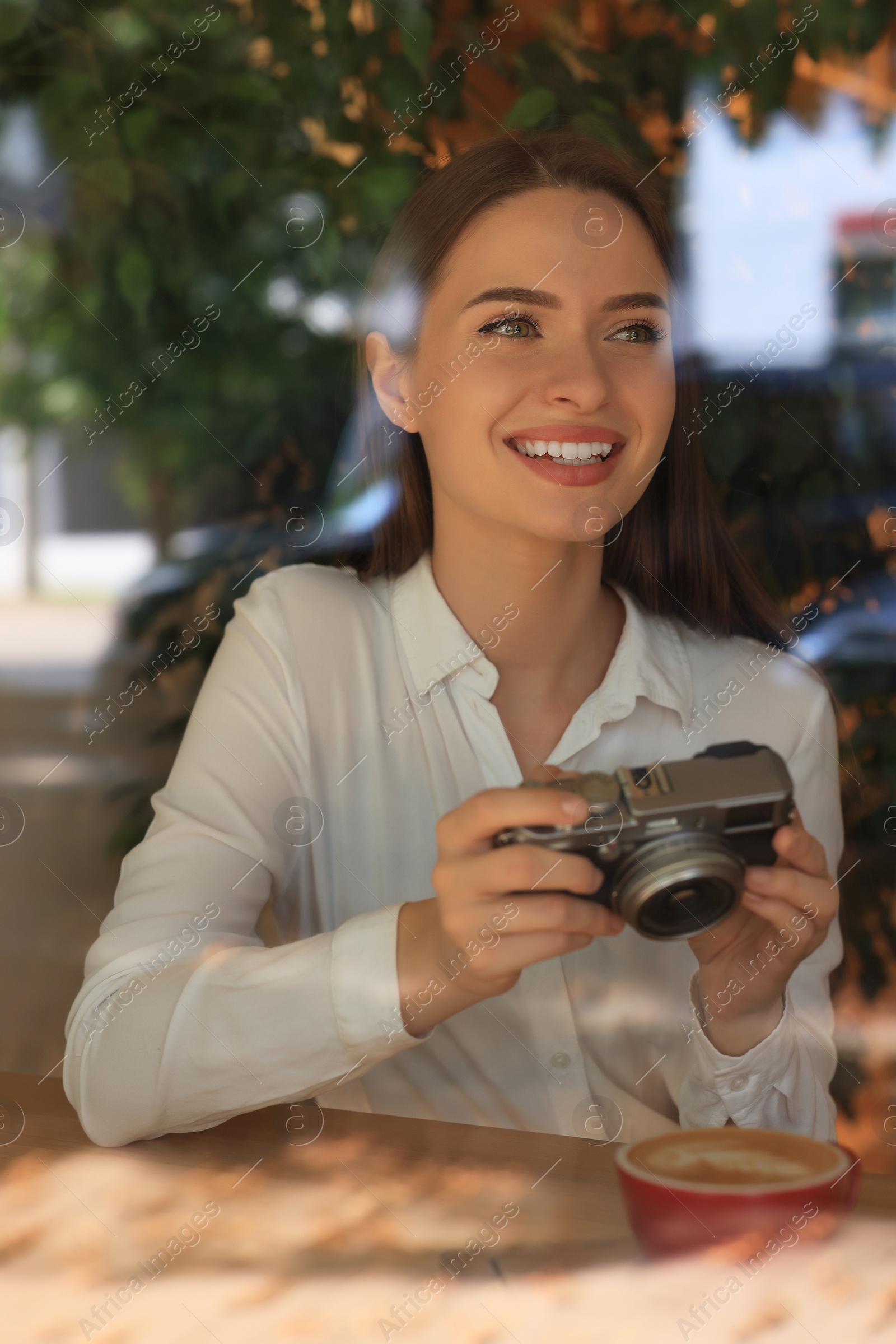 Photo of Young woman with camera and cup of coffee at cafe, view through window. Creative hobby