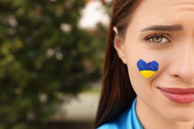 Photo of Sad young woman with drawing of Ukrainian flag on face outdoors, closeup. Space for text
