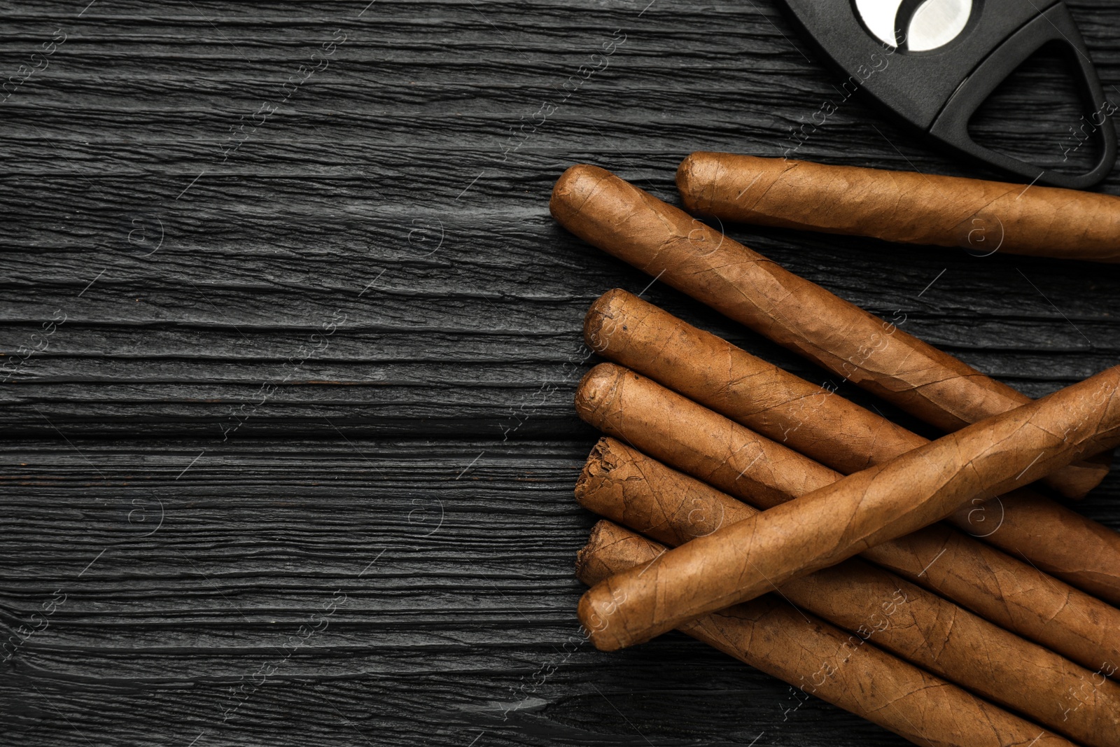 Photo of Cigars and guillotine cutter on black wooden table, flat lay. Space for text