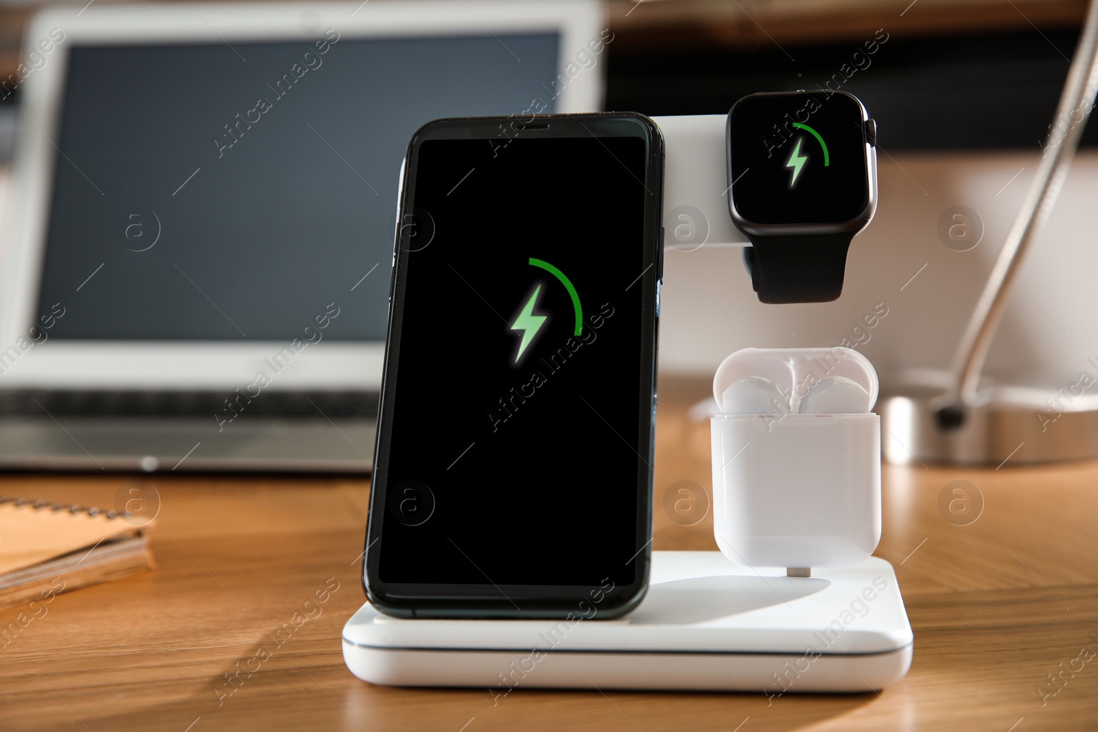 Photo of Mobile phone, earphones and smartwatch charging with wireless pad on wooden desk, closeup. Modern workplace device