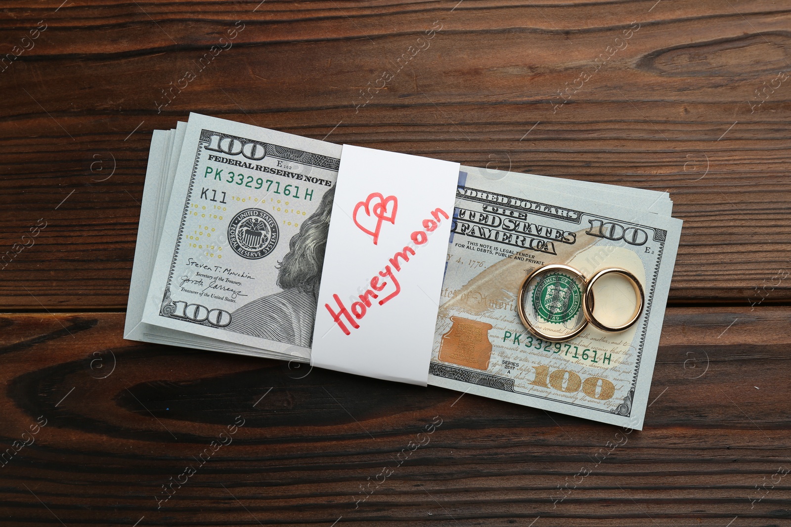 Photo of Stack of money with word Honeymoon and two golden rings on wooden table, top view