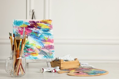 Photo of Different brushes, easel with abstract picture and paints on white wooden table. Space for text