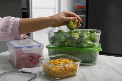 Woman putting green broccoli into glass container at white marble table in kitchen, closeup. Food storage