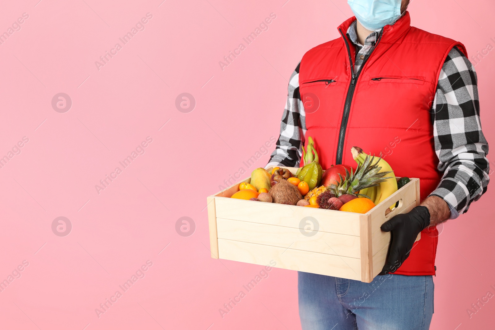 Photo of Courier holding crate with assortment of exotic fruits on pink background, closeup. Space for text