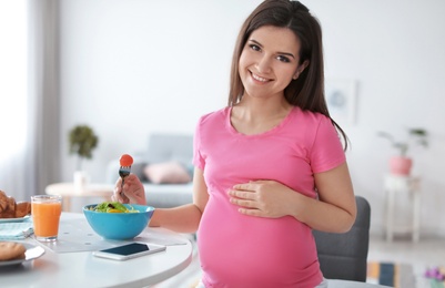 Photo of Young pregnant woman eating vegetable salad at table in kitchen