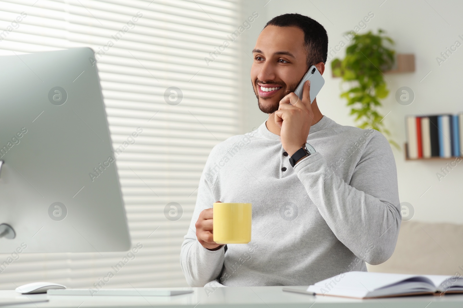 Photo of Young man talking on smartphone while working with computer at desk in room. Home office