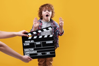 Photo of Emotional cute boy performing while second assistant camera holding clapperboard on orange background. Little actor