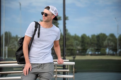 Photo of Handsome young man with stylish sunglasses and backpack near building outdoors, space for text