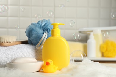 Baby cosmetic products, bath duck, brush and towel on white table against soap bubbles