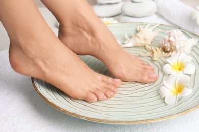 Woman soaking her feet in plate with water, flowers and seashells on white towel, closeup. Spa treatment