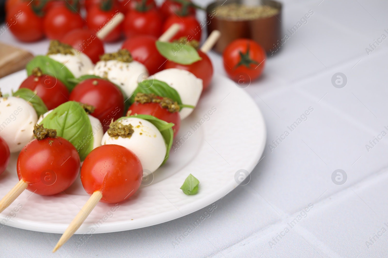 Photo of Caprese skewers with tomatoes, mozzarella balls, basil and pesto sauce on white tiled table, closeup. Space for text