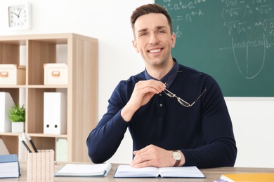 Photo of Young male teacher with book sitting at table in classroom