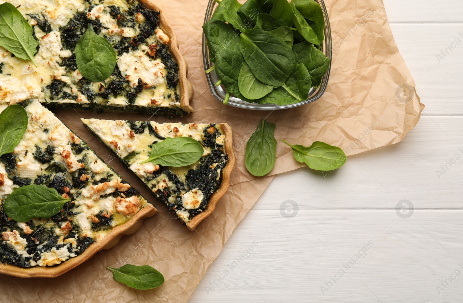 Photo of Delicious homemade quiche and fresh spinach leaves on white wooden table, flat lay