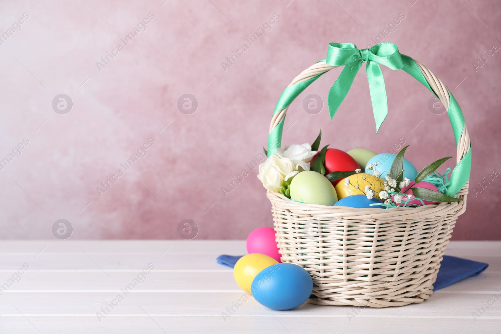 Photo of Wicker basket with bright painted Easter eggs and flowers on white wooden table against pink background. Space for text