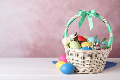 Wicker basket with bright painted Easter eggs and flowers on white wooden table against pink background. Space for text