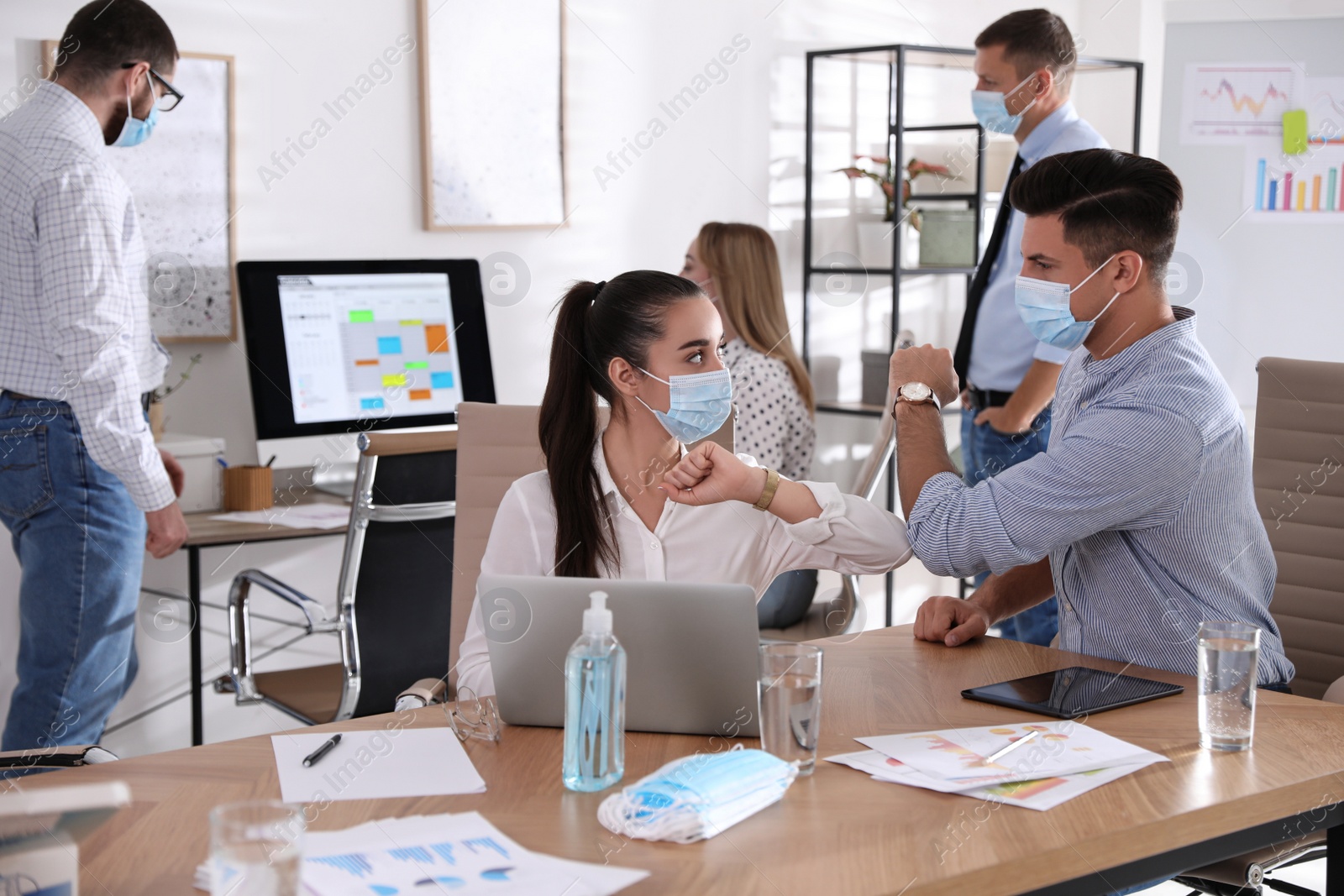 Photo of Coworkers with protective masks making elbow bump in office. Informal greeting during COVID-19 pandemic