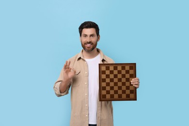 Smiling man with chessboard and showing OK gesture on light blue background