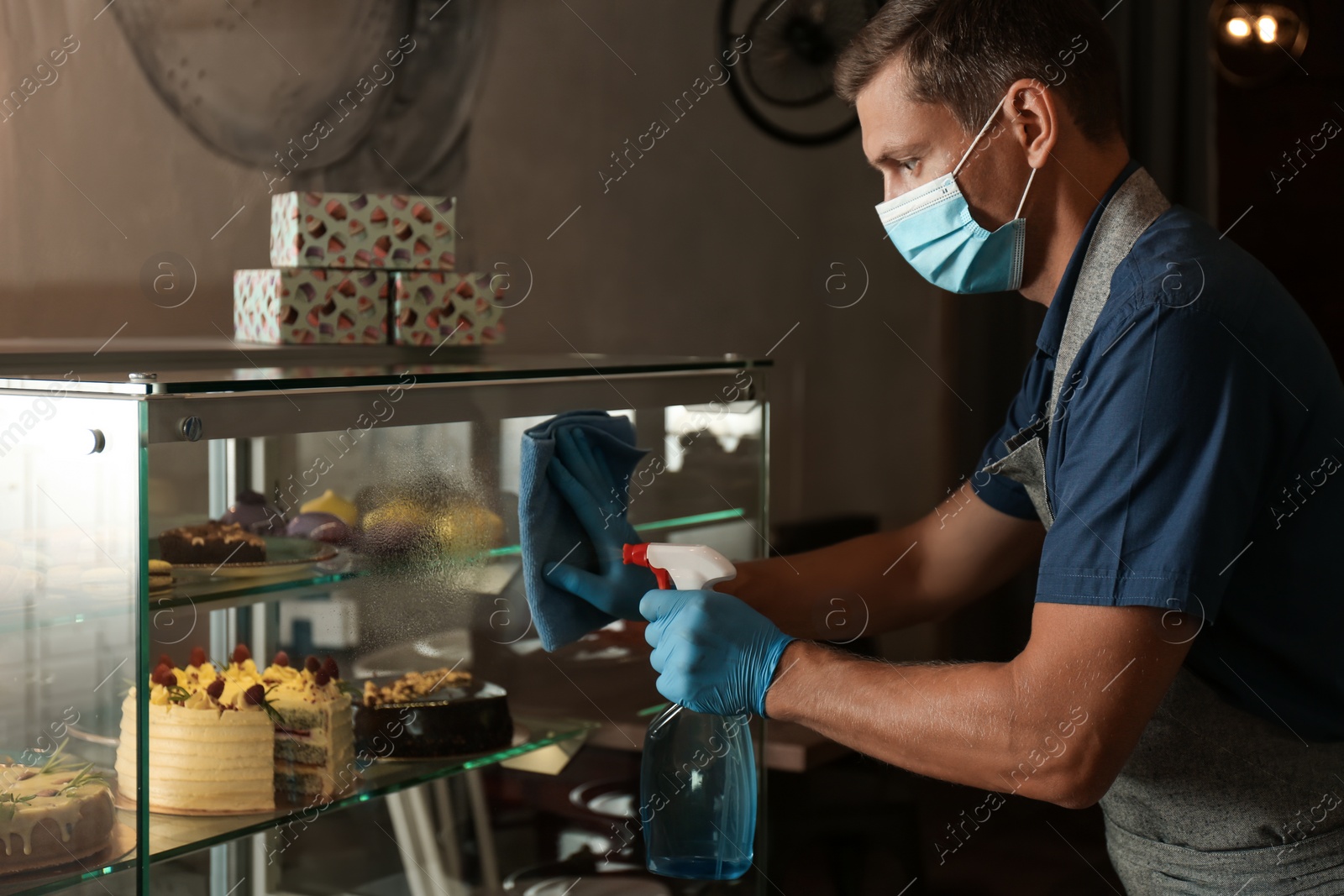 Photo of Worker in mask and gloves disinfecting dessert showcase at cafe