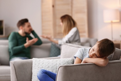 Little unhappy boy sitting in armchair while parents arguing at home