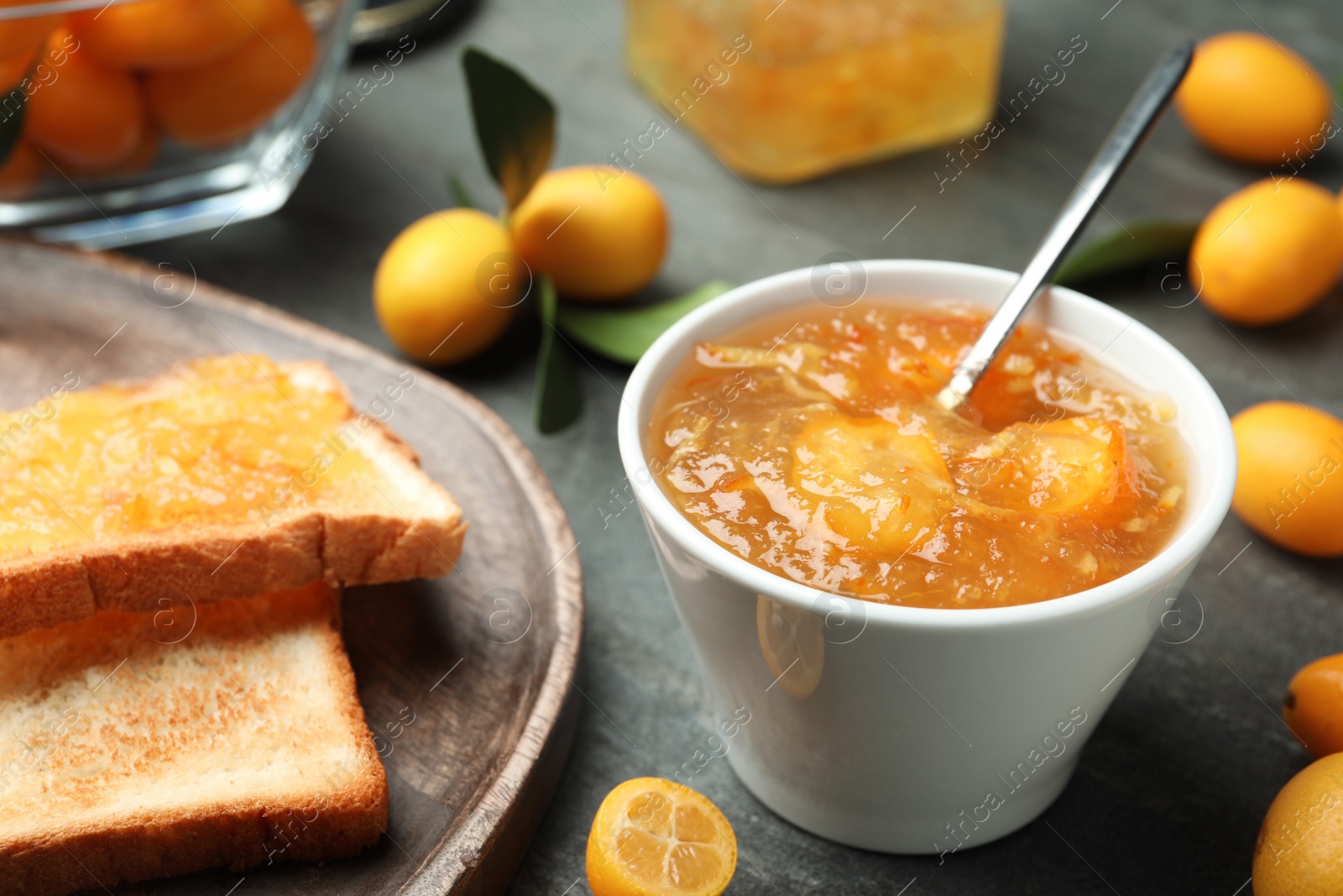 Photo of Delicious kumquat jam in bowl, toasts and fresh fruits on grey table, closeup