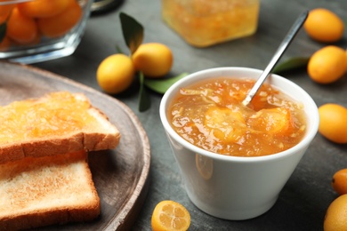 Delicious kumquat jam in bowl, toasts and fresh fruits on grey table, closeup