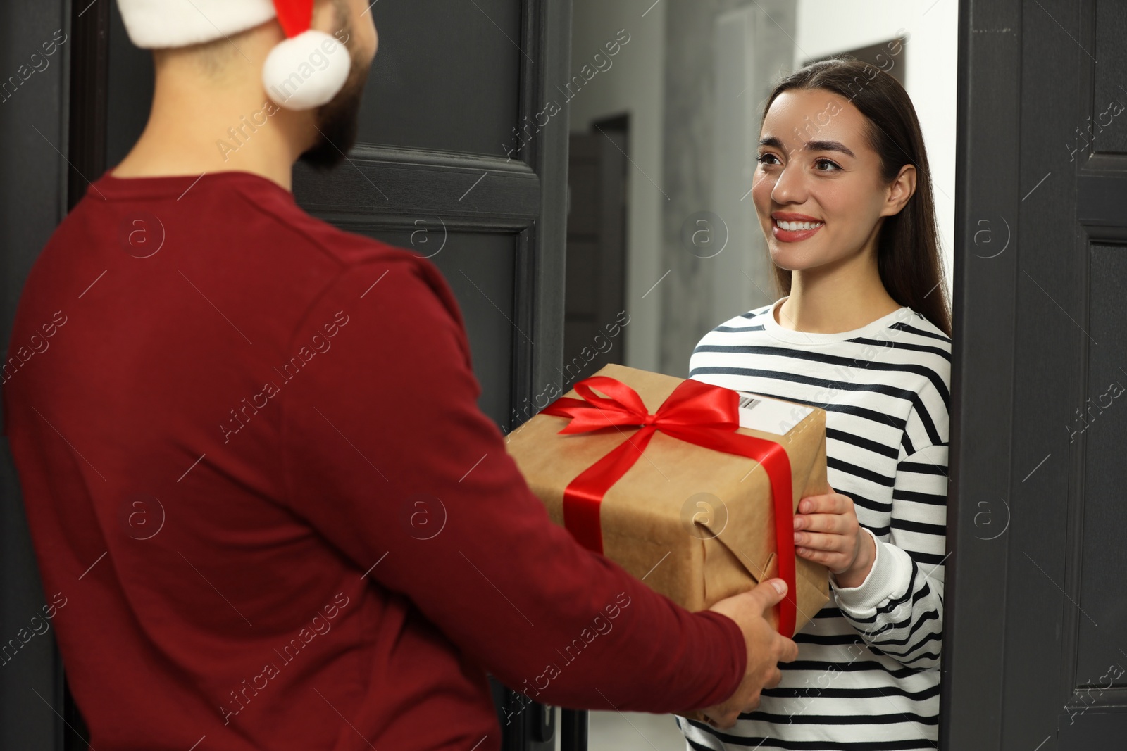 Photo of Courier in Santa hat giving young woman Christmas gift box indoors. Sending present by mail