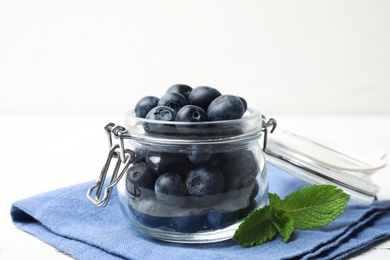 Glass jar of tasty fresh blueberries, mint leaves and fabric on white wooden table