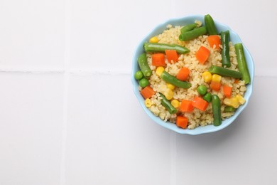 Photo of Delicious bulgur with vegetables in bowl on white tiled table, top view. Space for text