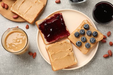 Photo of Tasty peanut butter sandwiches with jam, fresh blueberries and peanuts on gray table, flat lay