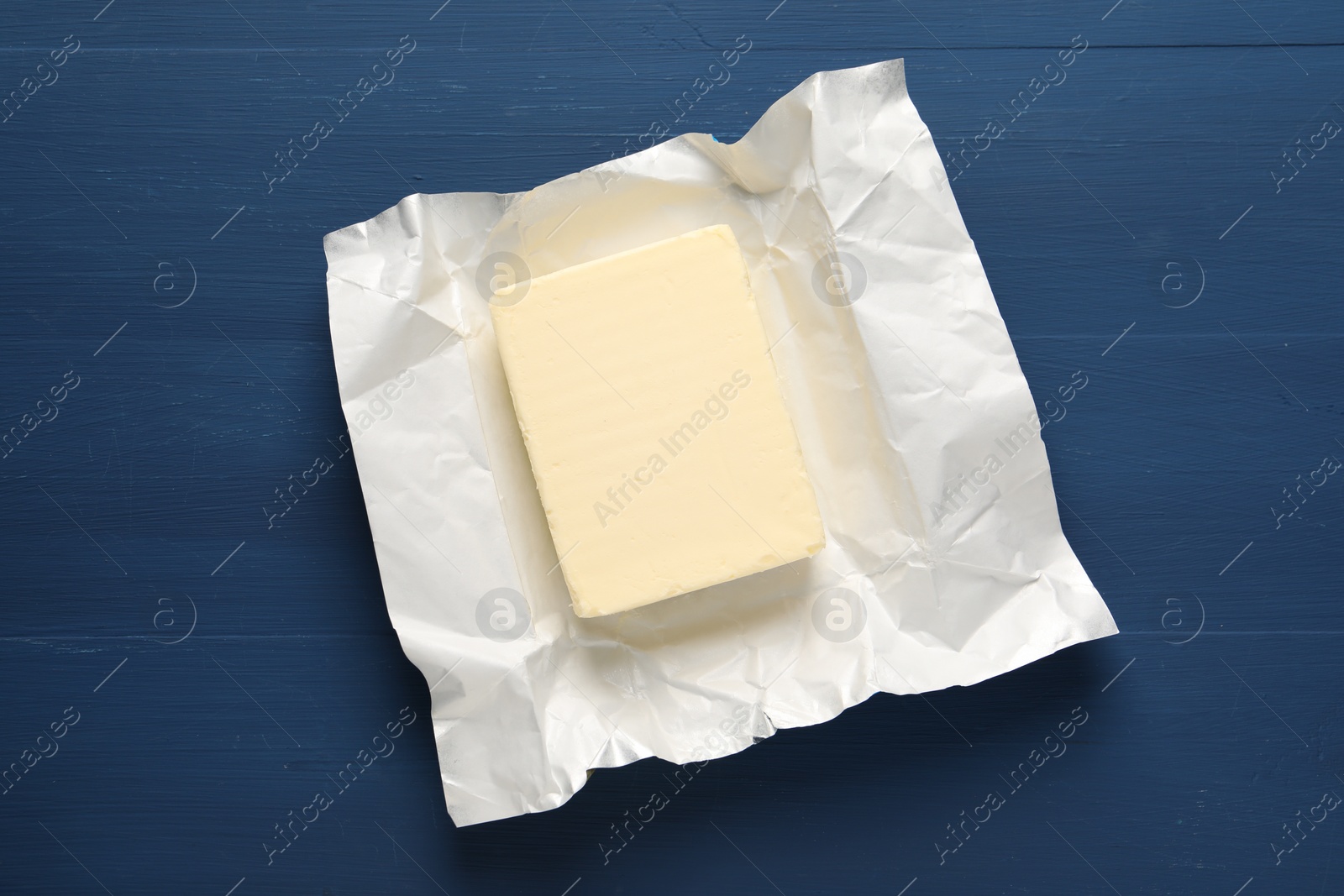 Photo of Block of tasty butter in open foil packaging on blue wooden table, top view