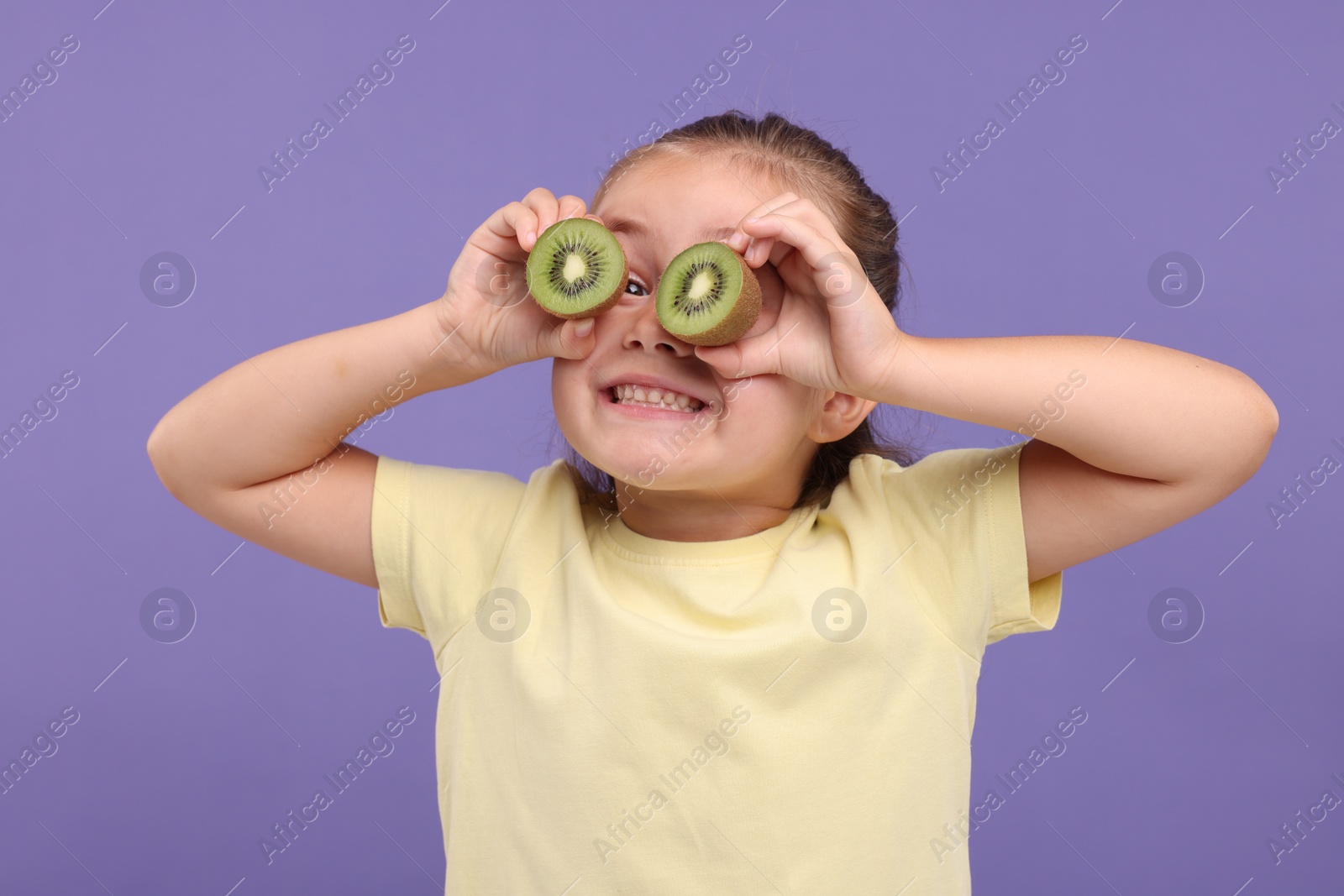 Photo of Smiling girl covering eyes with halves of fresh kiwi on violet background