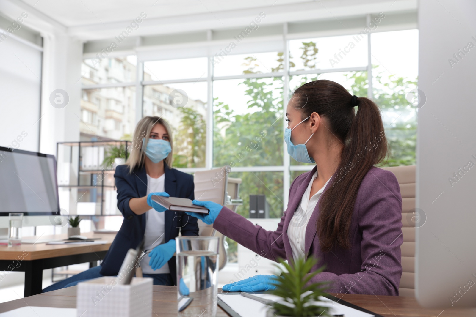 Photo of Office employee in mask and gloves giving notebook to her colleague at workplace