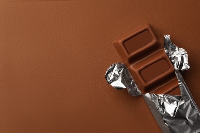Delicious chocolate bar wrapped in foil on brown background, top view. Space for text