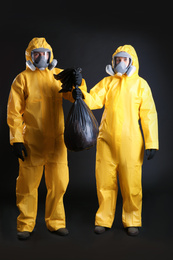 Photo of Man and woman in chemical protective suits holding trash bag on black background. Virus research