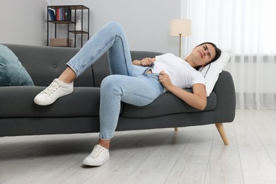 Young woman struggling to squeeze into tight jeans while lying on sofa at home