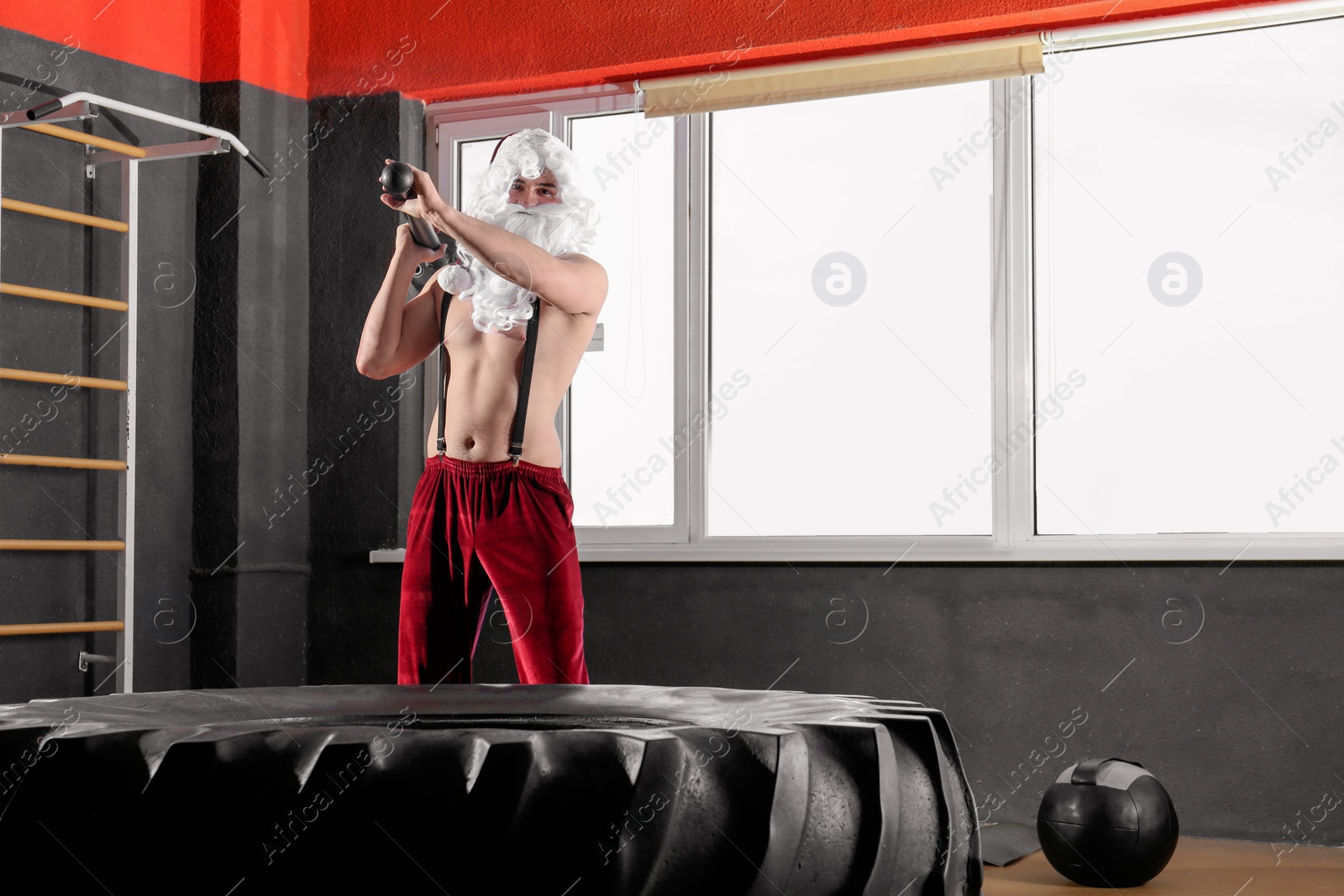 Photo of Young Santa Claus hitting heavy tire with hammer in modern gym