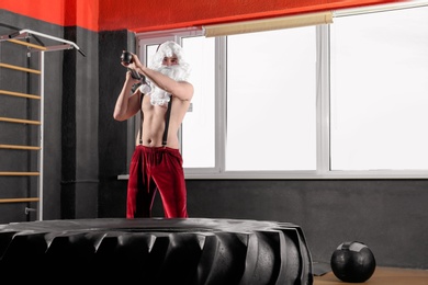 Photo of Young Santa Claus hitting heavy tire with hammer in modern gym