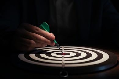 Business targeting concept. Man with dart aiming at dartboard at table in darkness, closeup