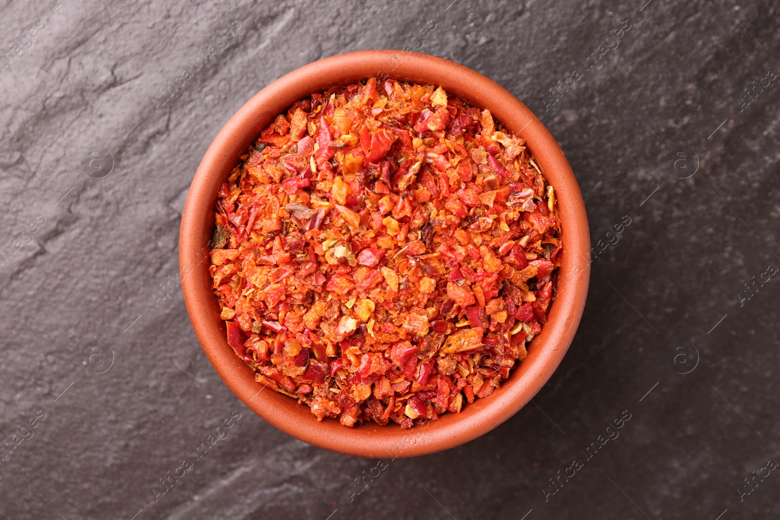 Photo of Chili pepper flakes in bowl on dark textured table, top view