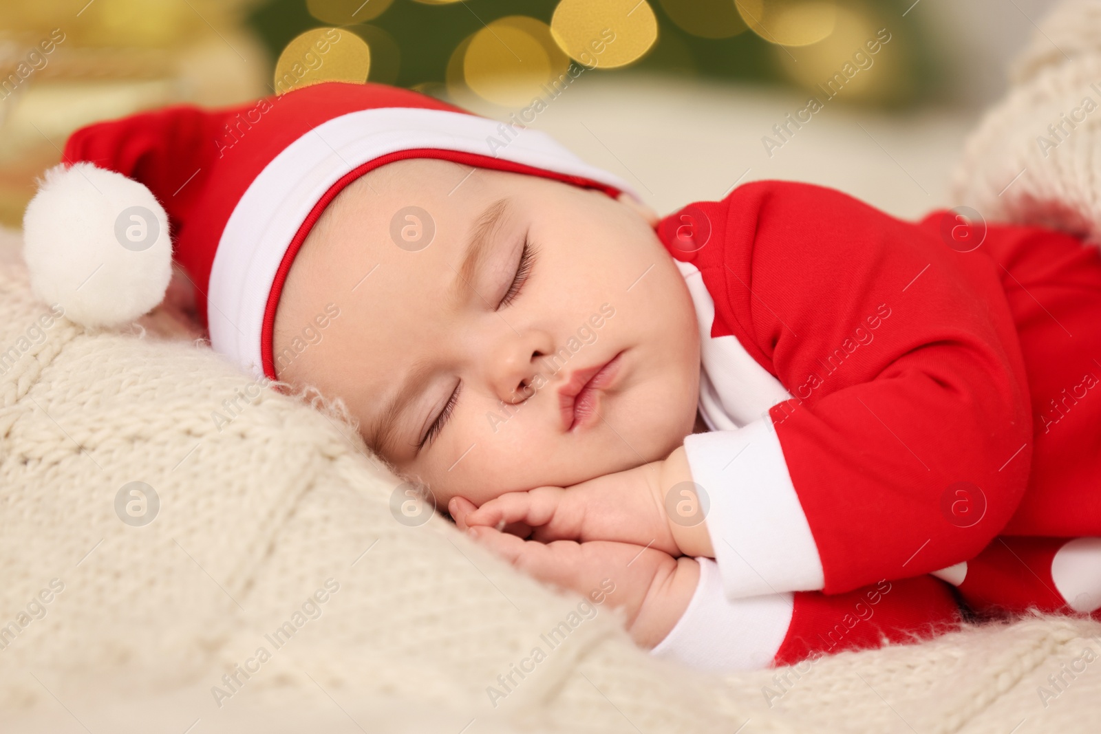 Photo of Cute baby in Christmas costume sleeping on knitted blanket against blurred festive lights. Winter holiday