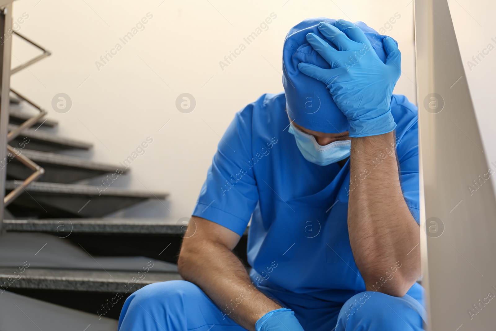 Photo of Exhausted doctor sitting on stairs indoors. Stress of health care workers during COVID-19 pandemic