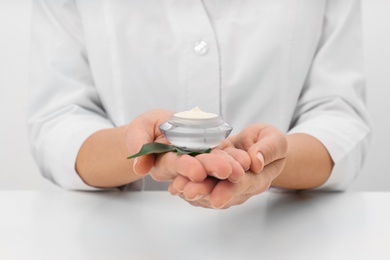 Photo of Female dermatologist holding jar of skin care product at table, closeup