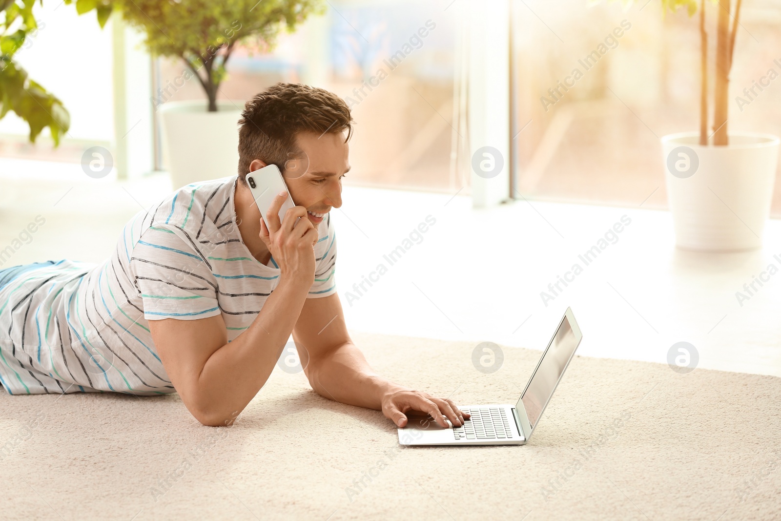 Photo of Handsome young man with mobile phone and laptop on cozy carpet at home